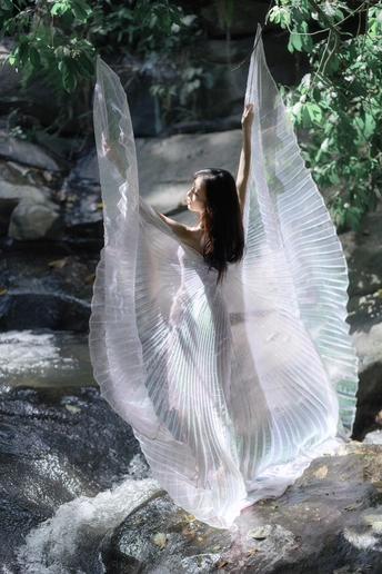 站在小溪或河里的岩石上的一个穿着带翅膀的白色连衣裙的年轻女子