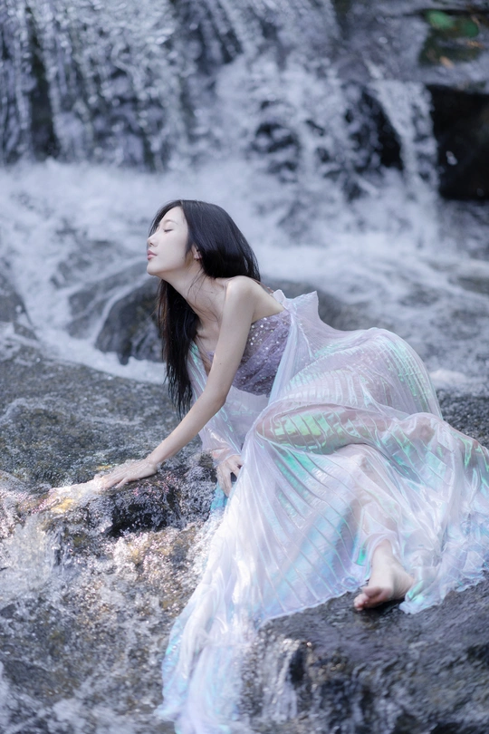 一位穿着蓝色连衣裙的年轻美女坐在瀑布边的岩石上
