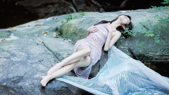 一个穿着蓝色连衣裙的漂亮年轻女子拿着一个塑料袋躺在溪流旁的岩石上