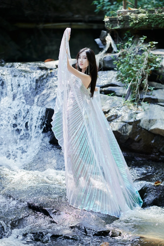 一位身穿白色长裙的美丽年轻女士站在以瀑布为背景的溪流中