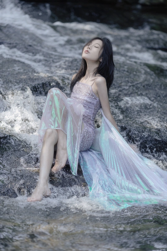 穿着蓝色连衣裙的美丽年轻女子坐在流动的水中的岩石上