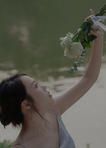 一位身穿白色连衣裙的年轻女子在水体附近将一束白玫瑰放在头上