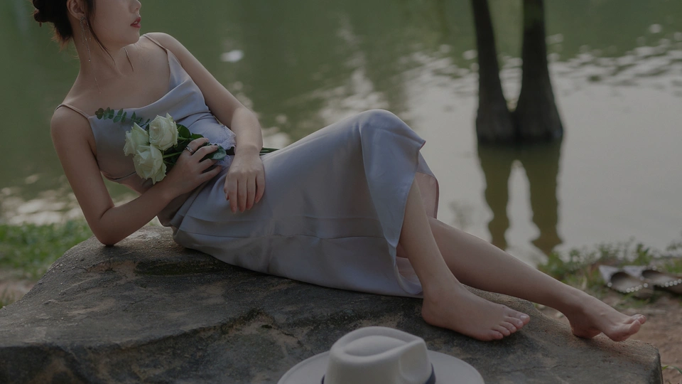 一个穿着连衣裙、戴着帽子的年轻女子坐在湖边的岩石上 手里拿着一杯咖啡、鲜花和一棵树