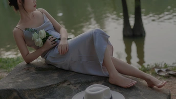 一个穿着连衣裙、戴着帽子的年轻女子坐在湖边的岩石上 手里拿着一杯咖啡、鲜花和一棵树