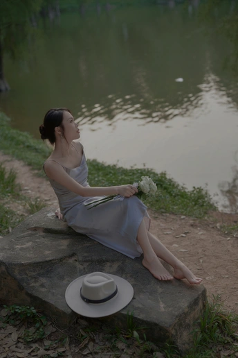 一个穿着连衣裙、戴着白色帽子的年轻女子坐在湖边或河边的岩石上 手里拿着一朵白花