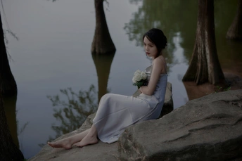 一个穿着白色连衣裙的年轻女子坐在湖边或河边的岩石上 背景是树和水