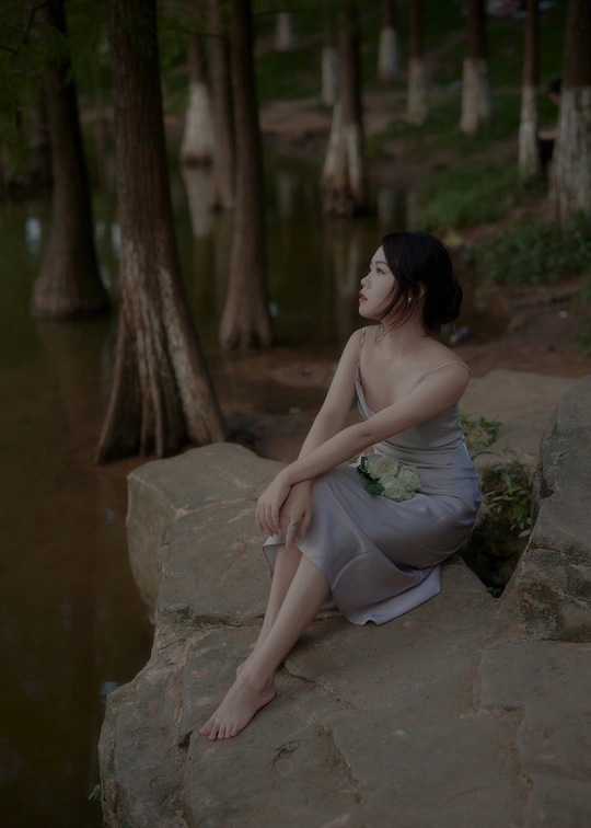 一个穿着连衣裙的年轻女子坐在水边和树旁的岩石上