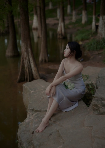 一个穿着连衣裙的年轻女子坐在水边和树旁的岩石上