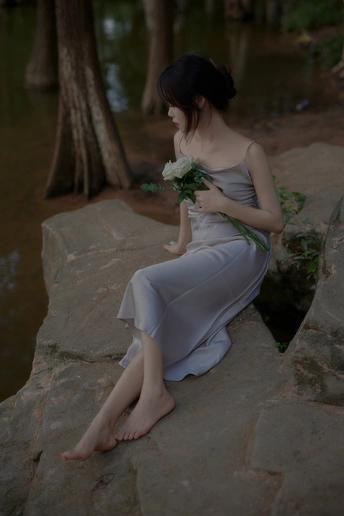 一位身穿白色连衣裙、手捧鲜花的年轻女子坐在水边的岩石上
