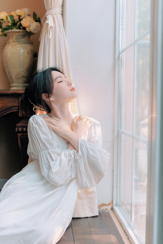 一个穿着白色连衣裙的年轻女子坐在地板上 凝视着房间里的一个花瓶。