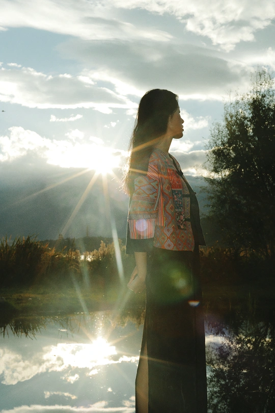 夕阳西下 一位女士站在湖边 阳光照耀在天空中 水面上波光粼粼。