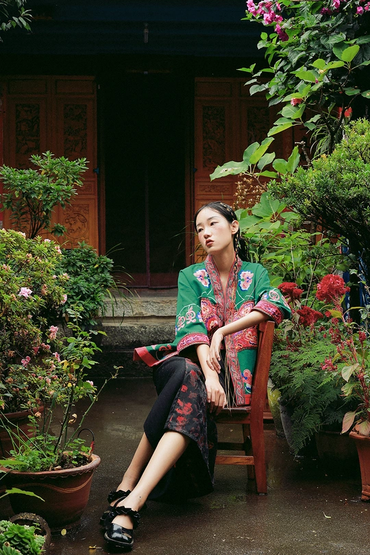 一个年轻女子坐在木凳上 前面是房子 院子里有植物和花。