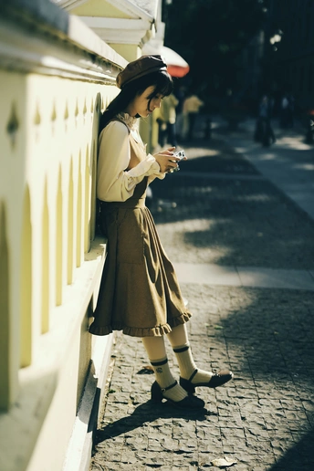 一位戴帽子穿裙子的年轻女子站在人行道上 倚靠在黄色墙壁上 正在看手机。