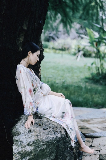 一个穿着连衣裙的年轻女子坐在树下的一块岩石上