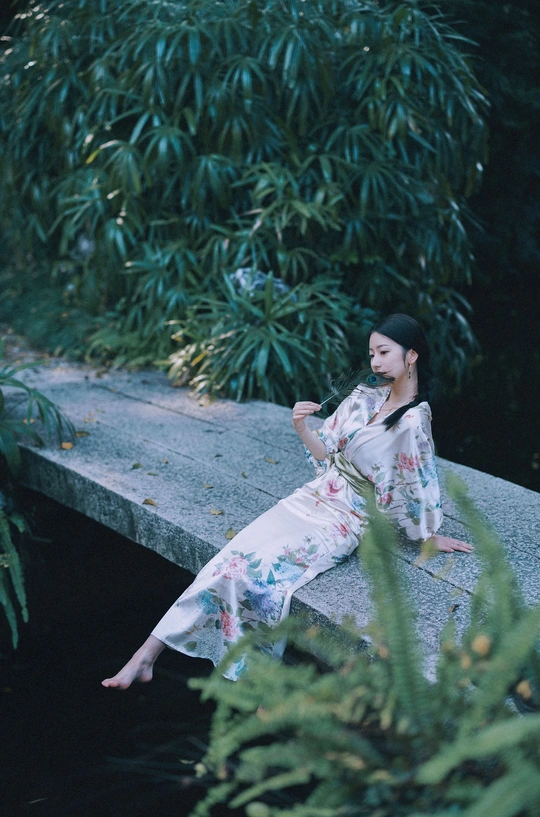 一个穿着连衣裙的年轻女子坐在桥上 桥下是流水 她手里拿着一把伞。