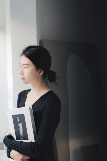 一个穿着黑色连衣裙的年轻女子拿着一本书站在窗户附近