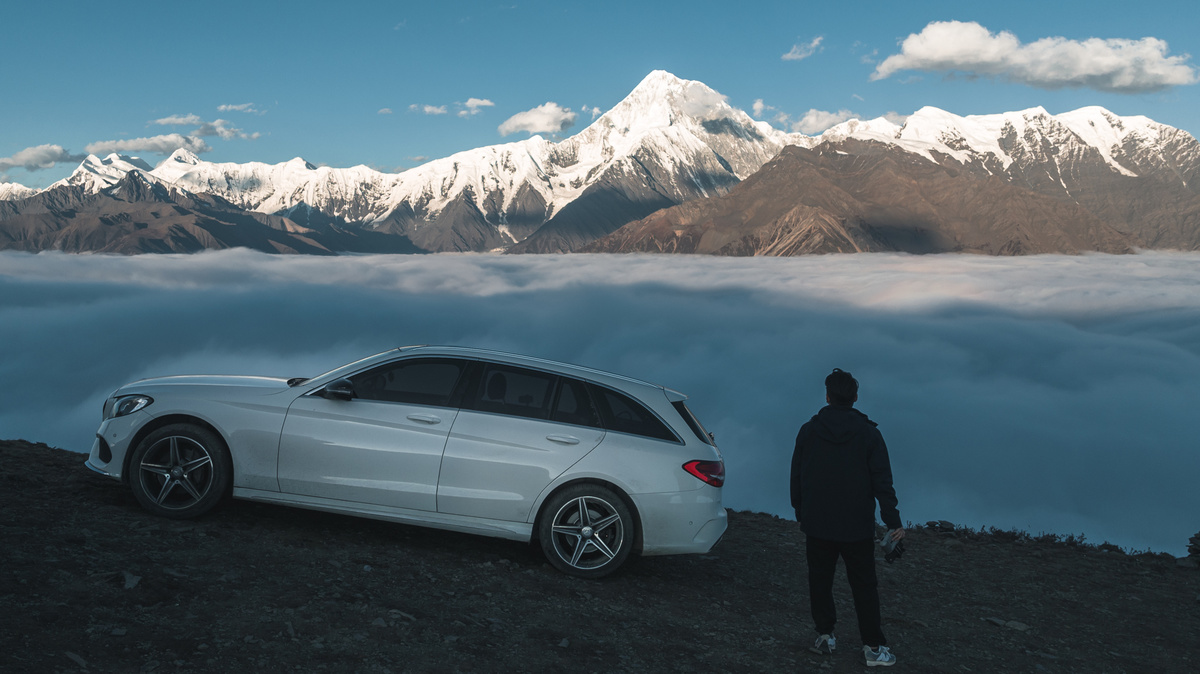 一辆白色汽车停在一个俯瞰山谷和云海以及湖泊的山坡上 一个男子站在汽车顶部。