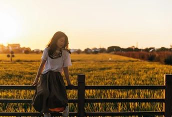 一个年轻女子穿着裙子站在日落时分的田地里的木质围墙上