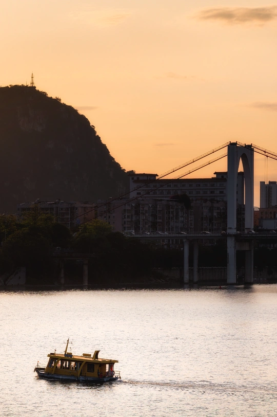 一艘小船在夕阳下漂浮在靠近城市和一座桥的大型水域上。