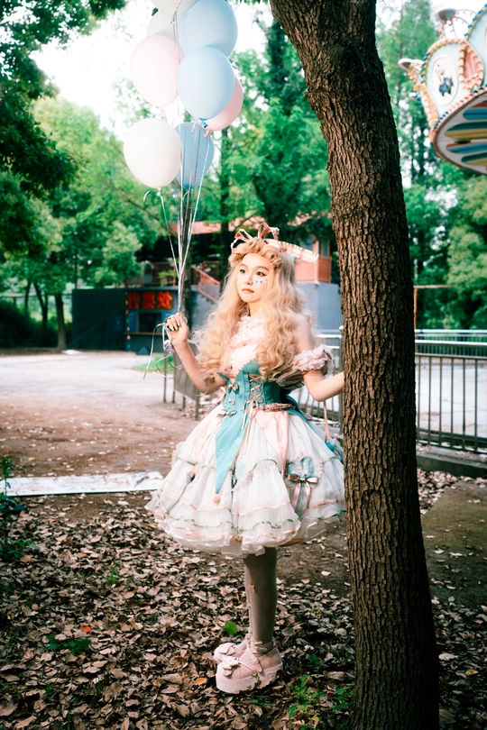 一个小女孩穿着仙女装 拿着气球站在公园里一棵树旁边。