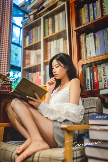 一个年轻女子坐在书架前的一把椅子上 正在阅读一本书 并看着一个白色的平板电脑上的笔记本。