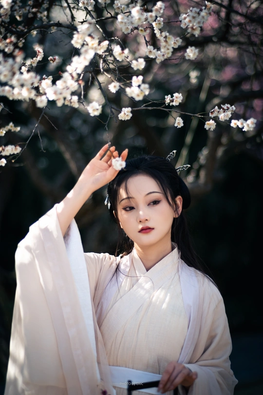 一位穿着传统白色连衣裙的美丽年轻女子站在樱花盛开的一棵树下。