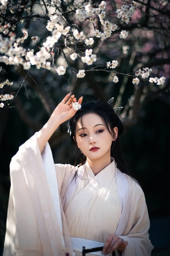 一位穿着传统白色连衣裙的美丽年轻女子站在樱花盛开的一棵树下。