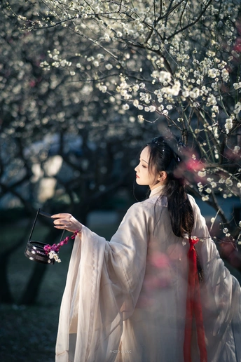 一位穿着传统白色和服的年轻女子 手里拿着花 站在盛开的花树下。