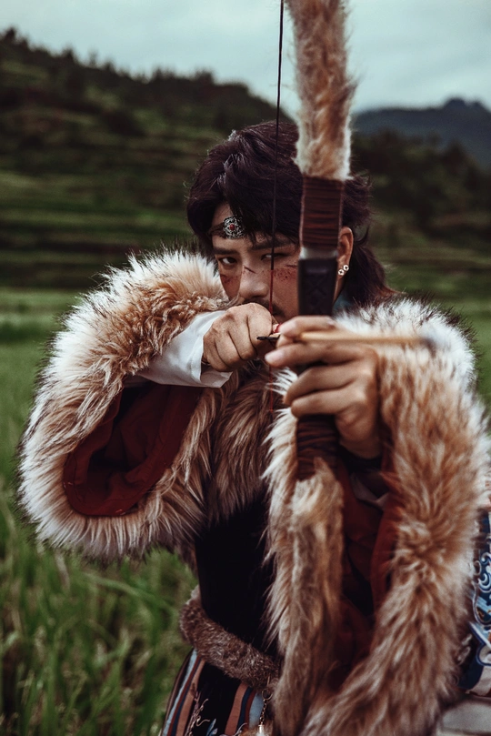 一名年轻男子穿着战士服装 手持弓箭和箭矢在田野中。