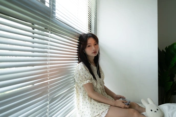 一个年轻女子坐在床上 前面有一个白色的毛绒玩具 窗户前有窗帘。