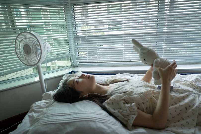 一位年轻女子躺在床上 前面有一个白色的毛绒玩具 窗前有窗帘和电风扇。