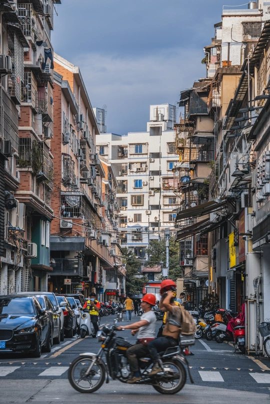 人们骑着滑板车和摩托车穿过一条建筑群排列的街道