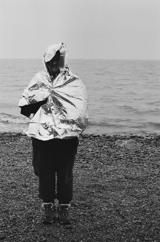 一位穿着雨衣带兜帽的女人站在水体岸边 背景中有一位男子在走动。