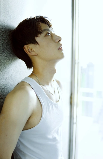 一个穿着白色背心的年轻人靠在墙上 正在透过窗户向外看。