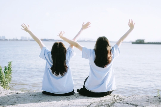 两个年轻女子坐在海滩上 手臂举过头顶 背景是一片水域。