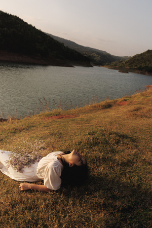 一个穿着白色连衣裙的年轻女子躺在草地上 她的头在水上方 靠近一个水体。