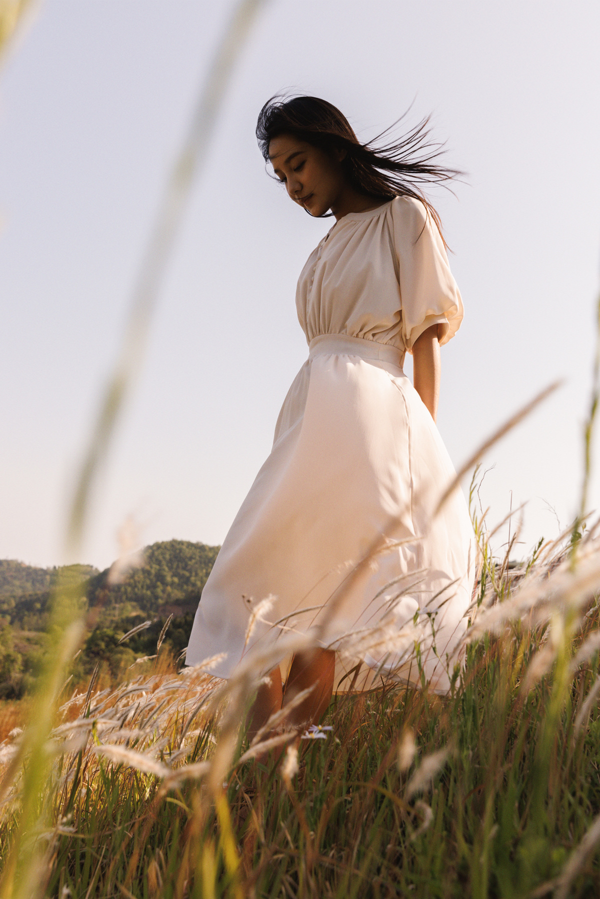一位穿着长白色连衣裙的年轻女子站在一片高草丛中。