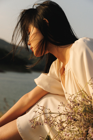 一位穿着白色连衣裙的年轻女子抱着一束花 坐在湖边。