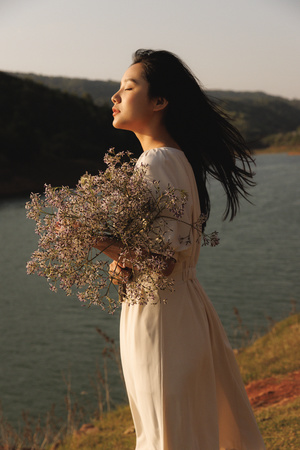 一位美丽的年轻女子穿着白色连衣裙 手里拿着一束花 站在悬崖上 俯瞰着一片水域。