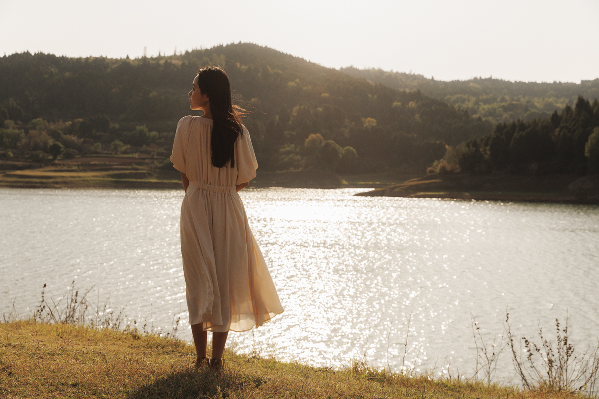 一位穿着白色连衣裙的年轻女子站在一个俯瞰湖泊的小山丘上。