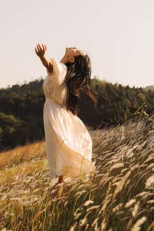 一个穿着长白色裙子的年轻女子站在一片高草地上 她的双臂向天空伸展。