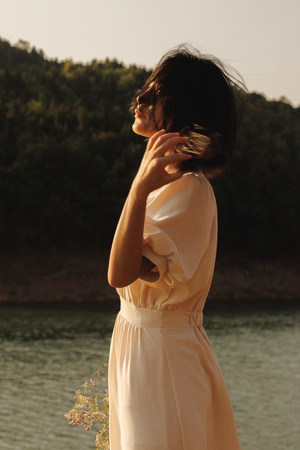 一位穿着白色连衣裙的年轻女子站在一水域旁 双手交叉在胸前。