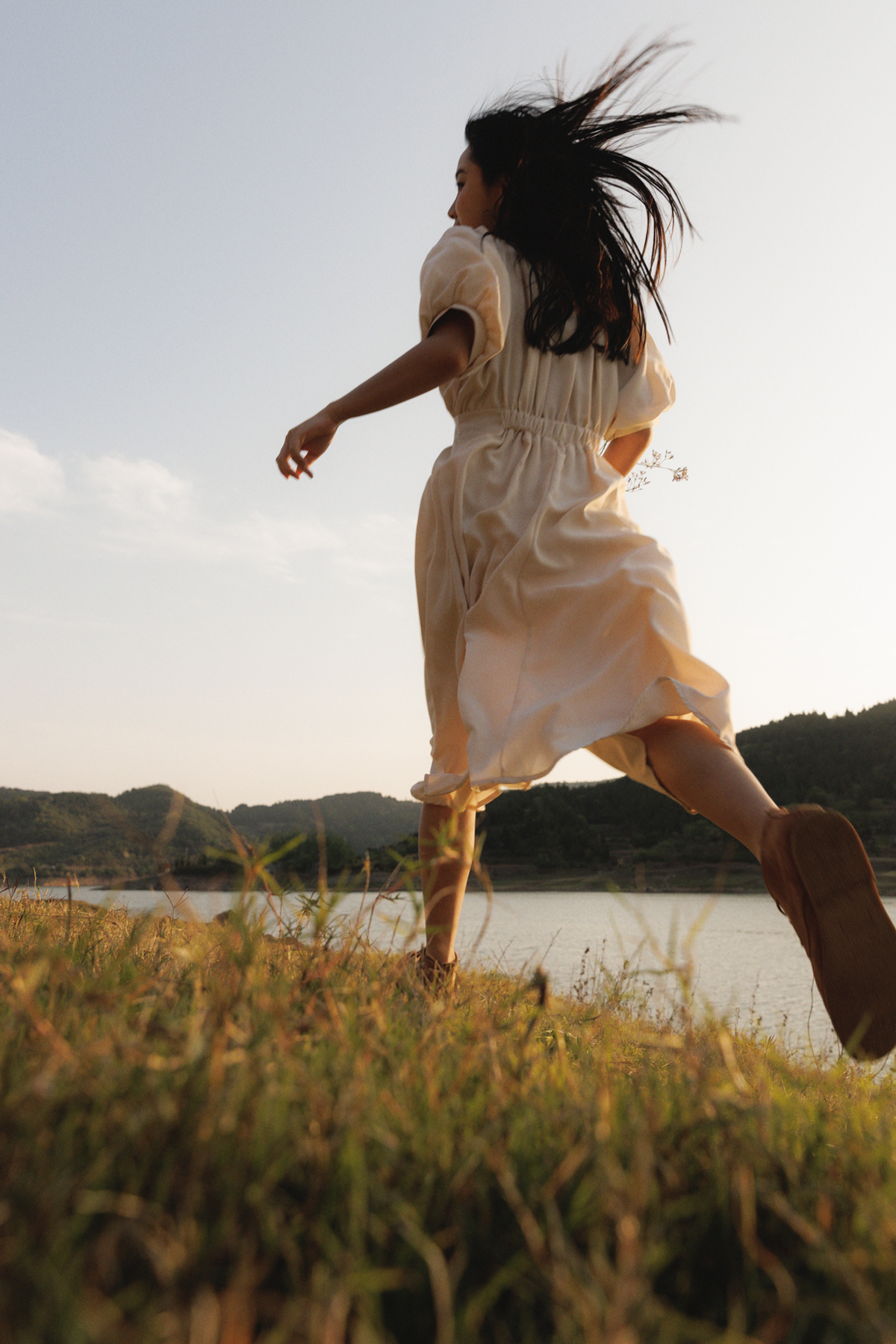 一位穿着白色连衣裙的年轻女子在湖边草地上跑过 手里拿着一把雨伞。