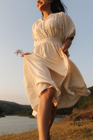 一位穿着白色连衣裙的年轻女子手持一朵花站在河边的一片田野上。