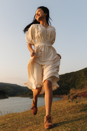 一位穿着白色连衣裙的年轻女子在湖边的山丘上跳跃 背景中有水。