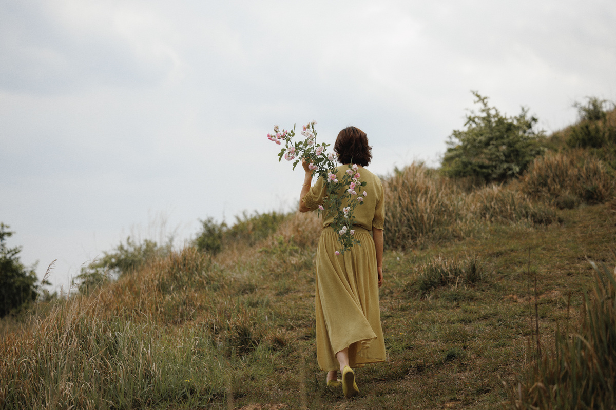 一位穿着黄色连衣裙 手拿一束花和一把伞 沿着山坡走的女人。