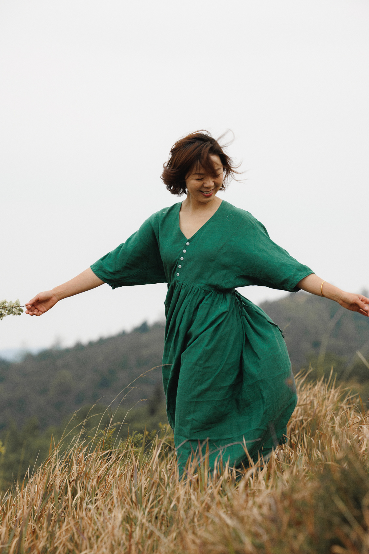 一位穿着绿色连衣裙的女士手里拿着一朵花 穿过一片草丛 走在田野里。