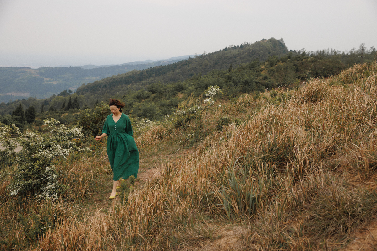 一位穿着绿色连衣裙的女士站在绿草如茵的山丘上 背景是山脉。