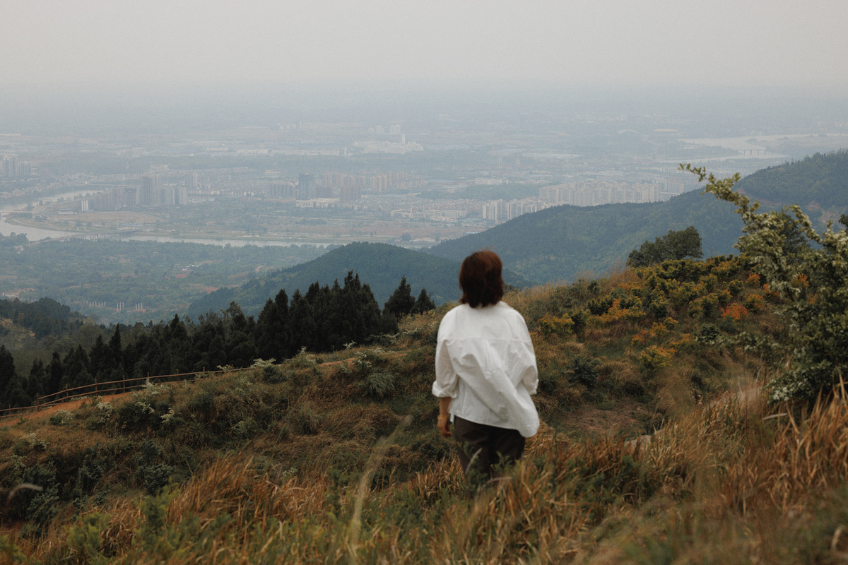 一位穿着白色衬衫的女士站在山顶上 俯瞰着山谷 远处有座城市。