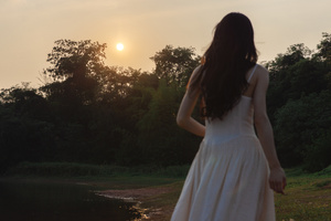 一位穿着白色连衣裙的年轻女子站在一个池塘前 当太阳落山时 水面上泛起一片金光。